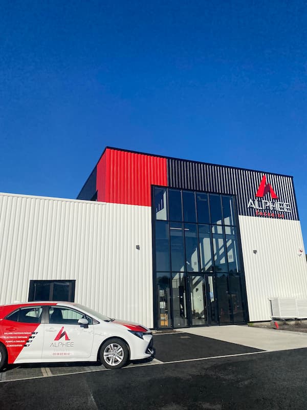 Façade moderne des nouveaux locaux d'Alphee Energies avec un design tricolore blanc, rouge et noir, une entrée vitrée sur toute la hauteur, et une voiture de société Alphee garée devant le bâtiment sous un ciel bleu vif.